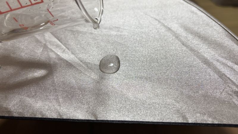 ユーロシルムのシルバー生地（防水スプレー前）に、ビーカーで水を一滴垂らした様子。まだ水玉の形を保っている。