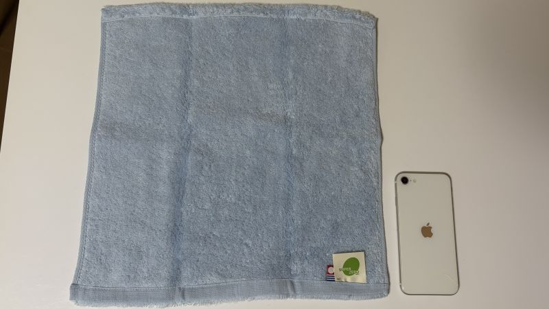 洗濯前のエアーかおる・わたのはなウォッシュタオルとiPhoneを並べた大きさ比較